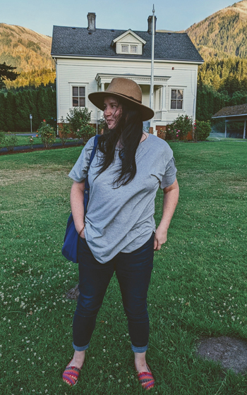 A woman standing in front of a house wearing a brown hat, grey shirt, denim jeans and multicolored shoes smiling while looking to the left over her shoulder while carrying a blue purse.