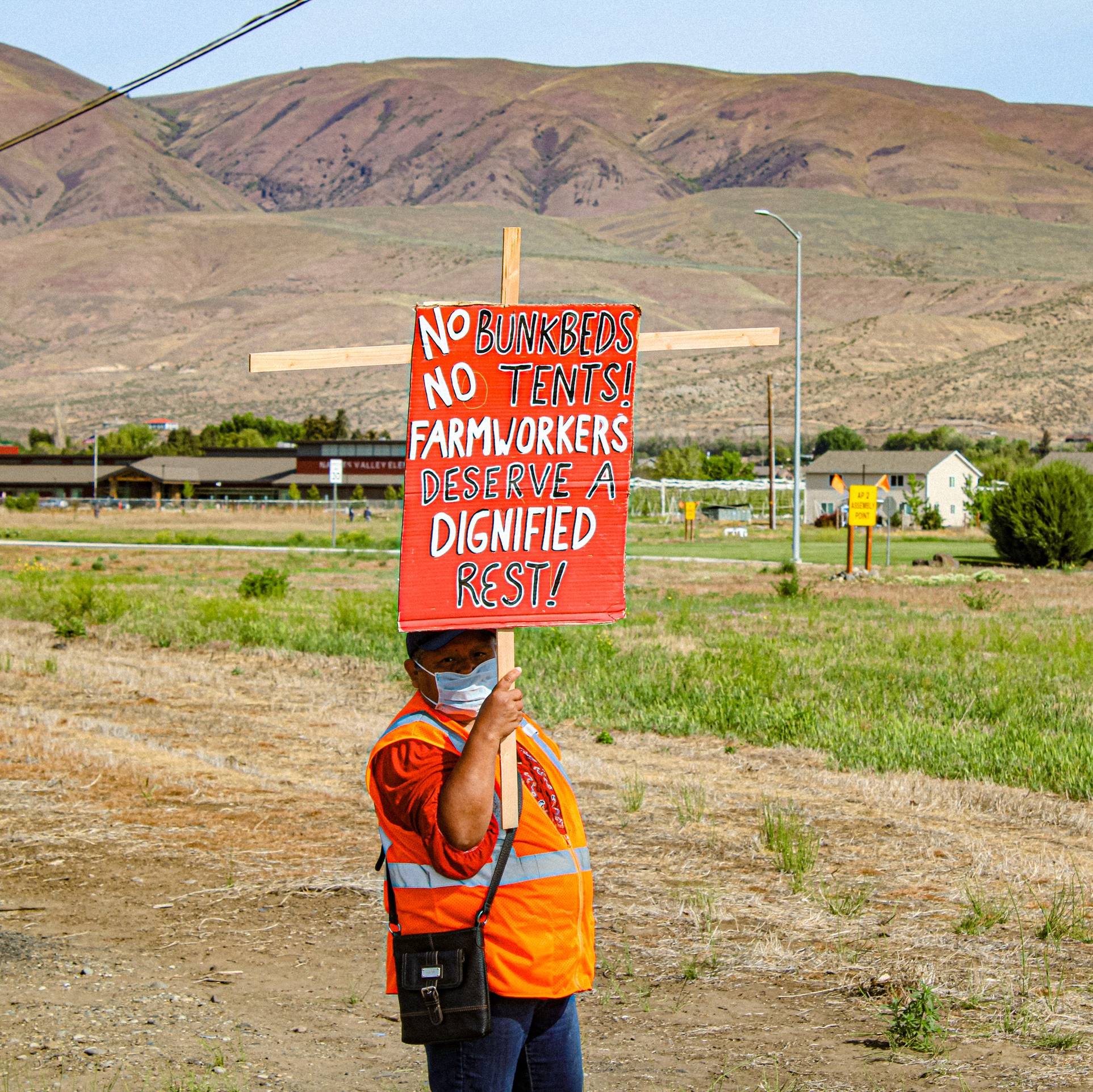 A person demonstrating at a farm holding a sign that reads “No bunkbeds, no tents, farmworkers deserve dignified rest!”