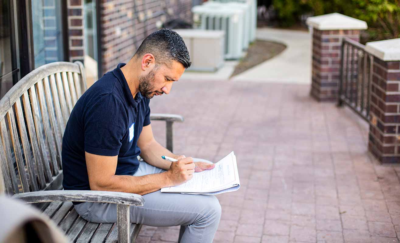 A man sits on a bench outdoors writing onto a piece of paper on his lap.