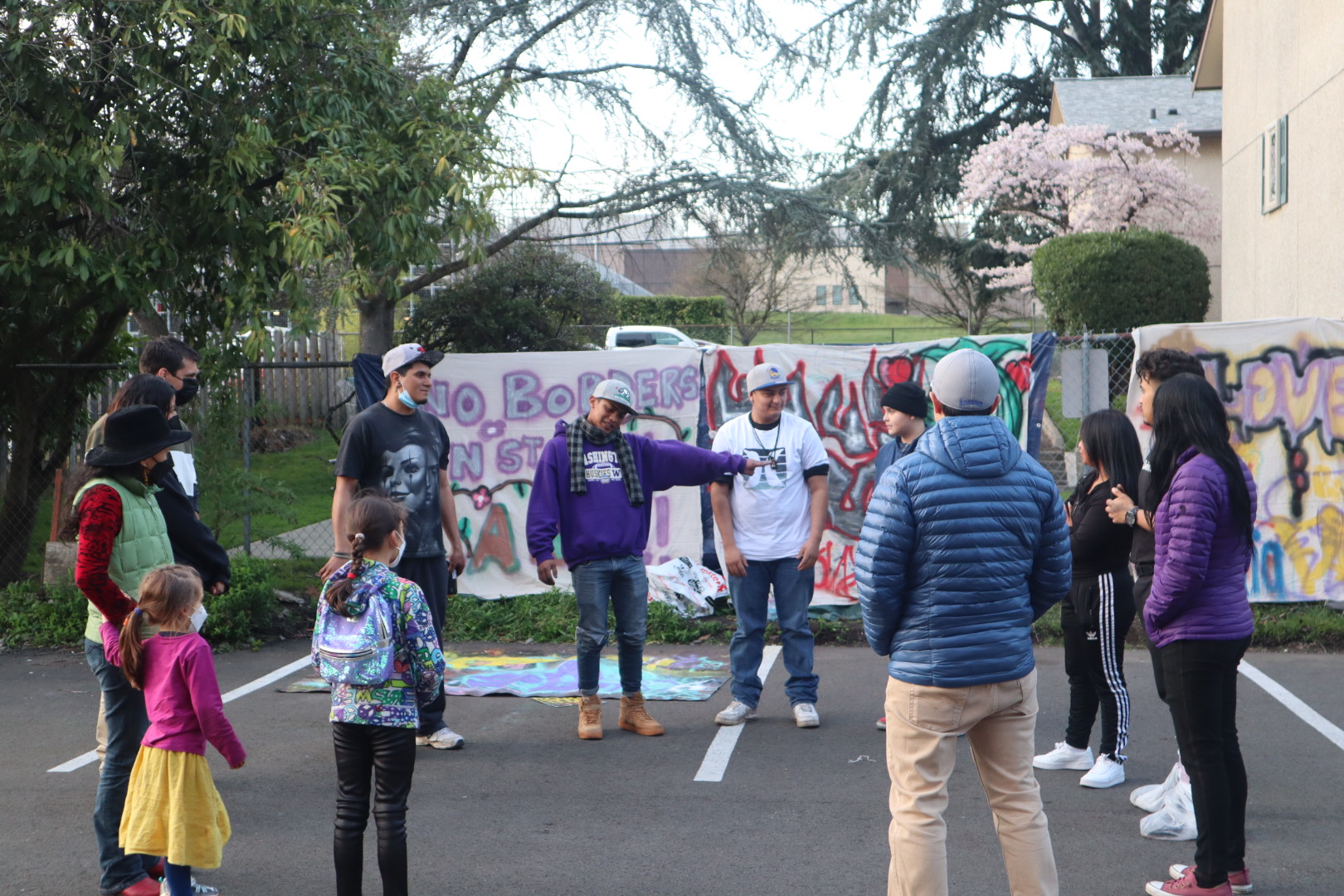 A group of community members, both children and adults, standing in the circle in a parking lot with hand-painted signs in the background.