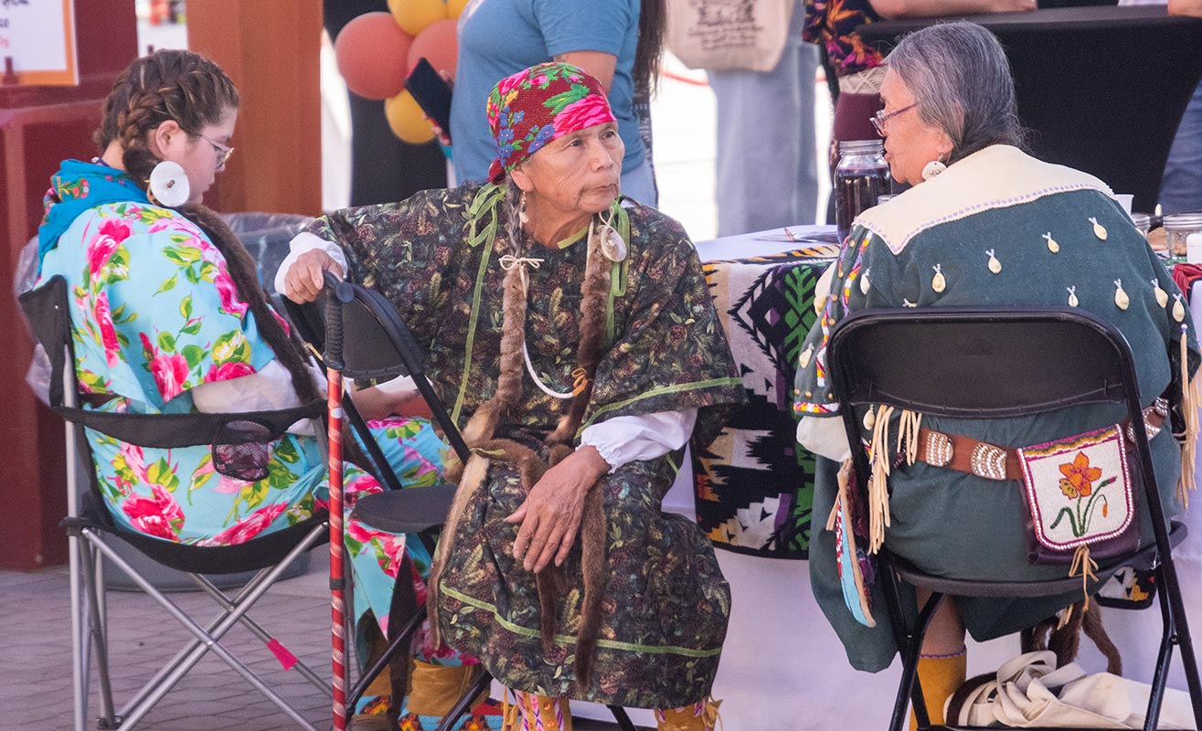 Two elders with the Yakama Nation are seated and talking with one another during an outdoor celebration.