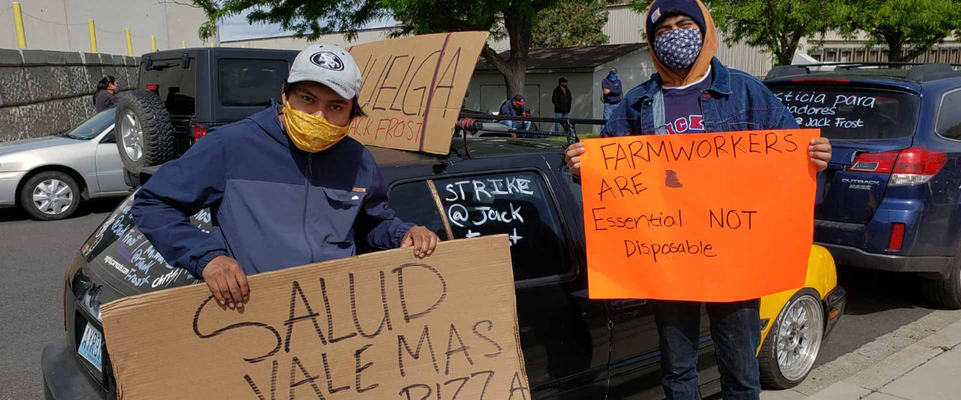 Two people wearing bandanas as face masks hold signs with one reading, “Farmworkers are essential not disposable.”