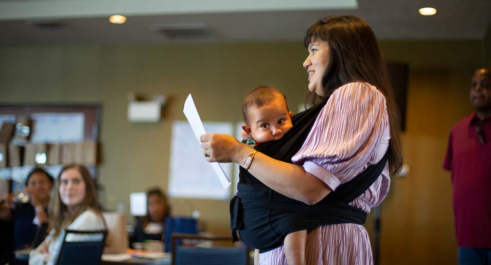  A woman stands to report out during a meeting while holding her baby in a harness.