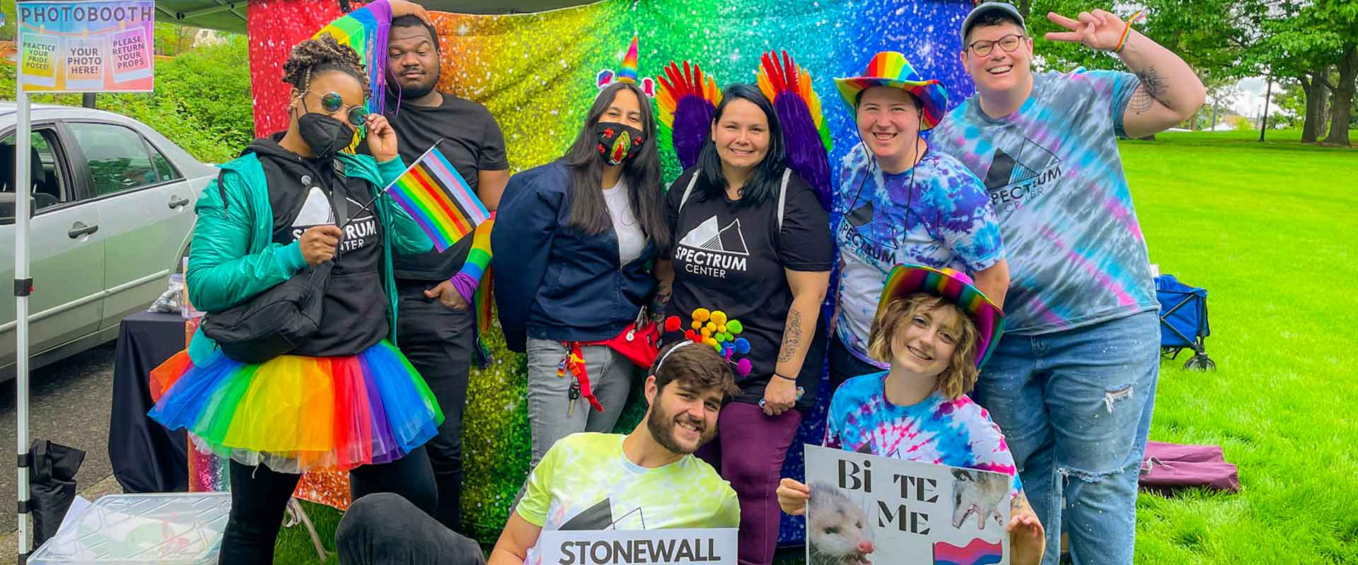 [Systems Change] A group of eight people in bright colors and holding signs posing for a photo in celebration of Pride.