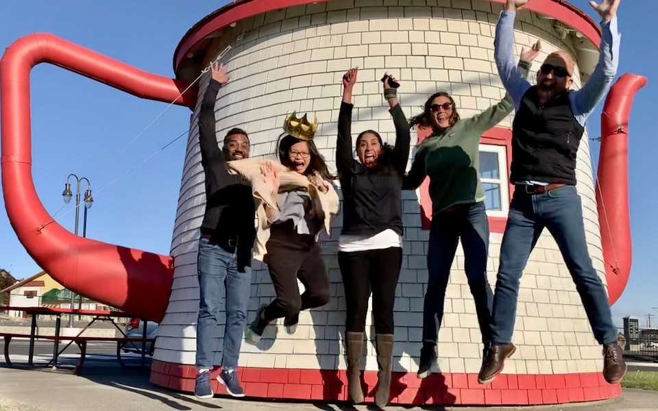 Five people jumping in front of a small building in the shape of a teapot.