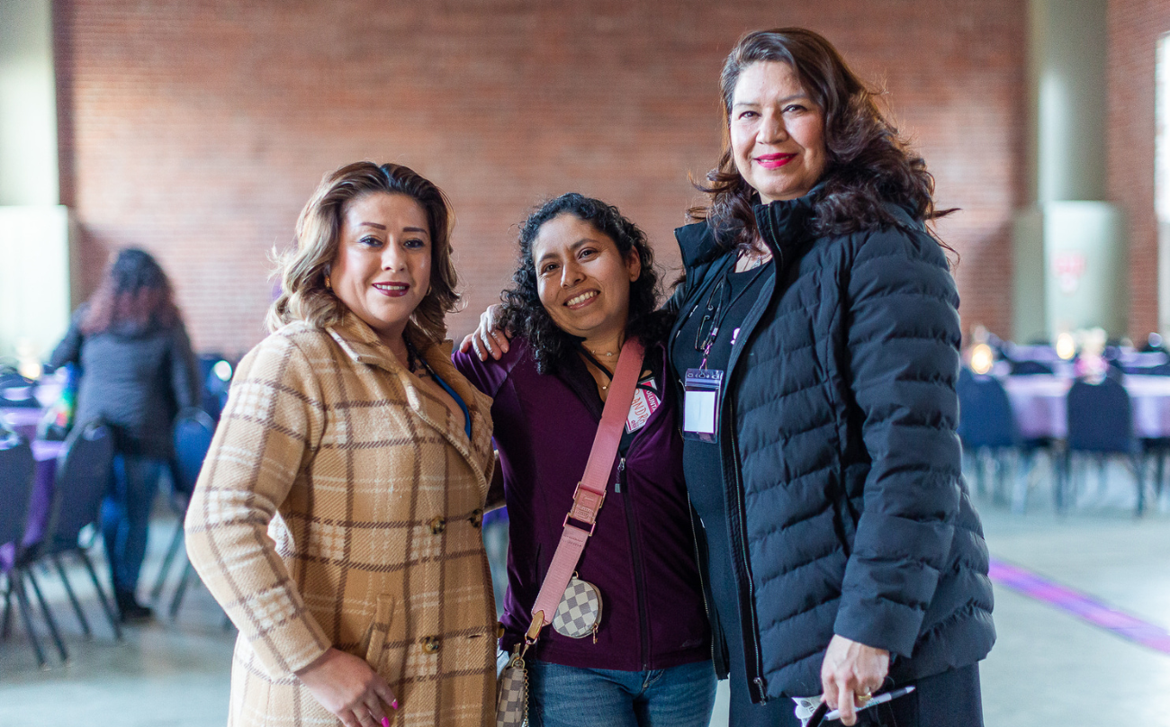 Tres mujeres latinas sonríen a la cámara en un acto comunitario.