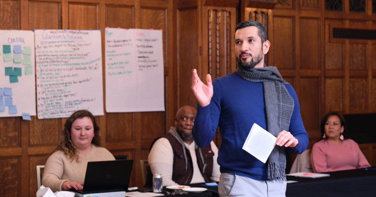 A man at the forefront wearing a blue sweater and gray scarf speaking with three people behind him